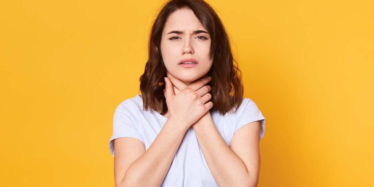 Studio shot of pleasant looking female with upset look, wears white casual t shirt, keeps both hands on neck, poses against yellow background. Beautiful woman feels bad because of sore throat.